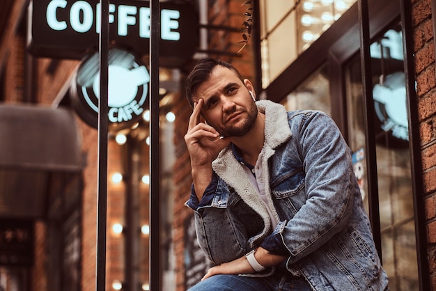 Портрет задумчивого стильного мужчины в джинсовой куртке с беспроводными наушниками, держащего кофе на вынос возле кафе.