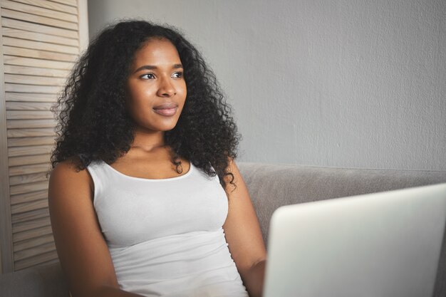 Portrait of pensive young African American female blogger uploading new video on her beauty vlog, sitting on sofa with electronic device on her lap, looking away with thoughtful dreamy smile