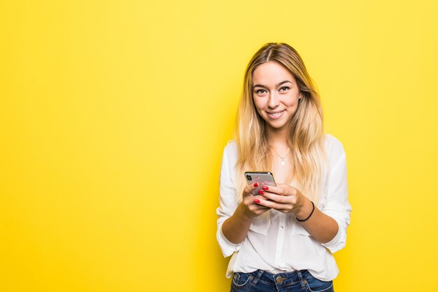 Портрет задумчивой молодой девушки, держащей мобильный телефон, стоя и глядя вдаль, изолированную над желтой стеной