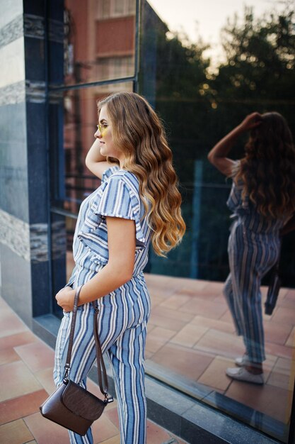 Portrait of a perfect young woman wearing striped overall and yellow sunglasses poses with her handbag on a balcony of a modern building in a town