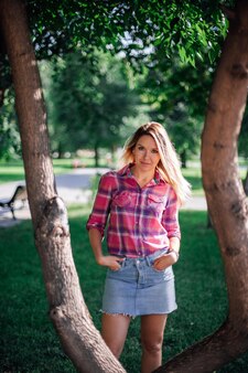 Portrait photo of adult shining blonde woman with hands in pockets standing between two trees in par...