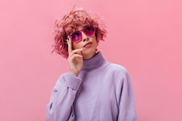 Портрет розоволосой кудрявой задумчивой женщины в фиолетовом свитере и солнцезащитных очках цвета фуксии позирует на изолированной стене