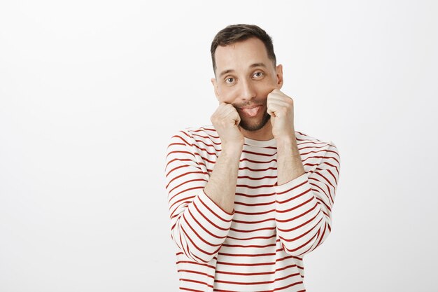 Portrait of playful childish Caucasian guy in striped clothes, pulling mouth with hands and sticking out tongue with indifferent bored expression