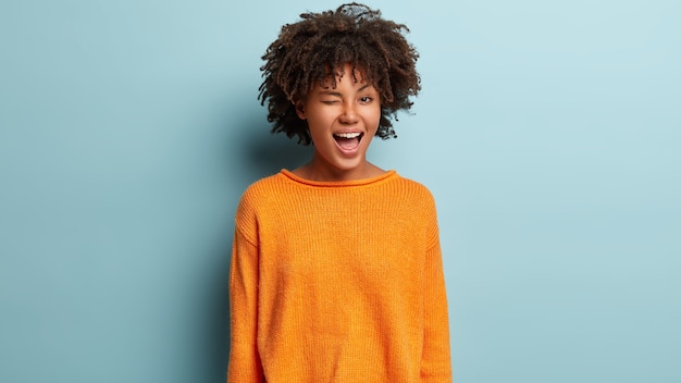 Free photo portrait of playful funny young woman with glad expression, blinks eye, flirts with boyfriend, expresses good emotions, wears orange jumper, isolated over blue wall.