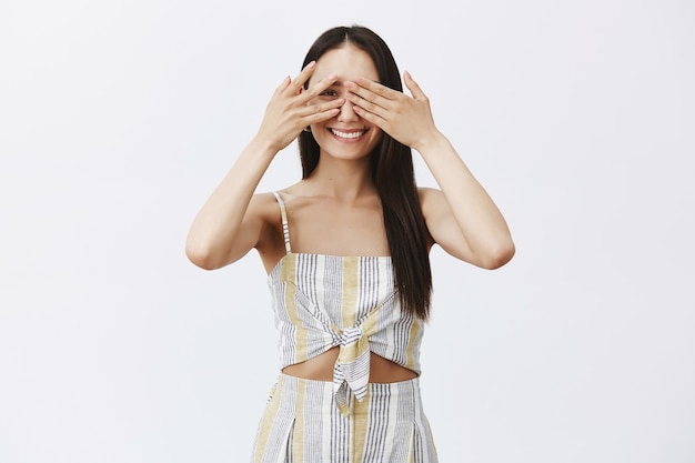 Portrait of playful happy attractive girl in matching clothes, covering eyes with palms and peeking through fingers joyfully, posing over gray wall