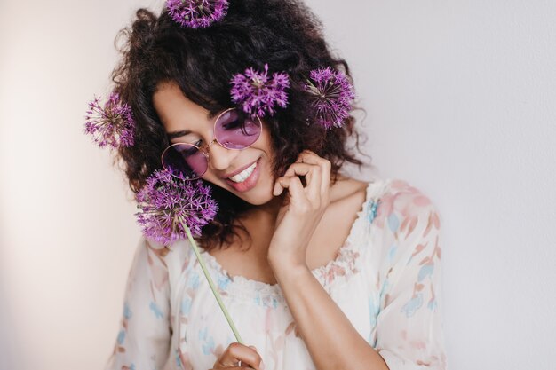 Portrait Of Pleasant African Girl Dreamy Posing. Black Young Woman With Purple Flowers In Hair Smiling During Photoshoot.