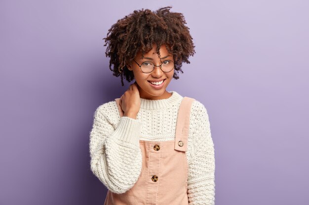 Portrait of pleasant looking dark skinned female with Afro haircut, keeps hand on neck, being in good mood