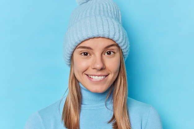 Portrait of pleasant looking European woman smiles gappily looks with interest at camera wears hat and casual turtleneck isolated over blue background. Cheerful teenage girl happy winter came