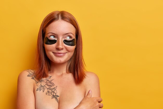 Portrait of pleasant looking redhead woman applies patches to reduce under eye puffiness and dark circles, has charming smile, stands shirtless against yellow wall. Beauty and rejuvenation