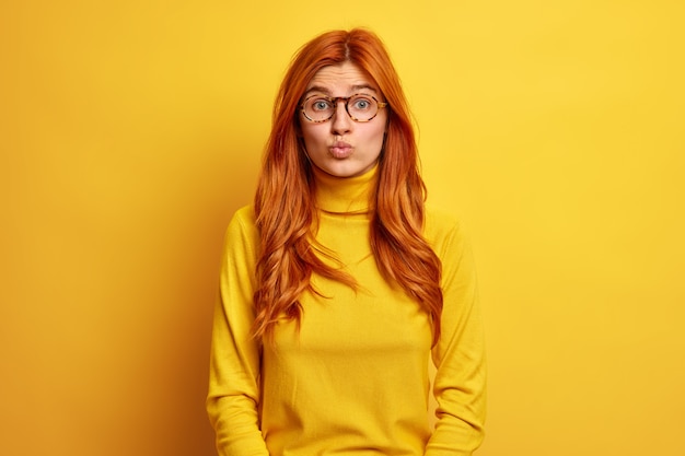 Portrait of pleasant looking redhead woman keeps lips rounded wants to kiss someone has romantic look wears optical glasses turtleneck.