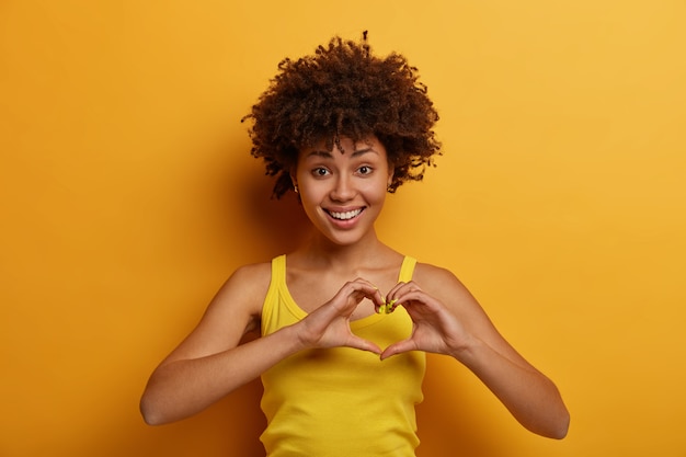 Free photo portrait of pleasant looking woman makes heart gesture, says be my valentine, smiles positively, expresses love and care, fell in love with someone, wears yellow shirt, stands indoor alone.