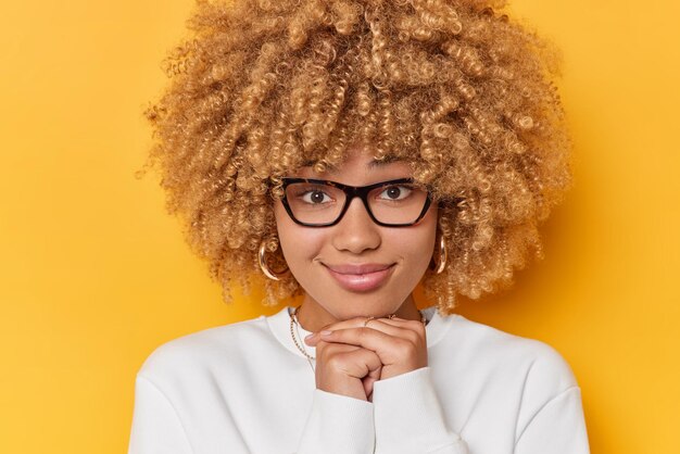 Portrait of pleasant looking woman with curly hair keeps hands under chin looks directly at camera listens information attentively wears spectacles and white jumper isolated over yellow background