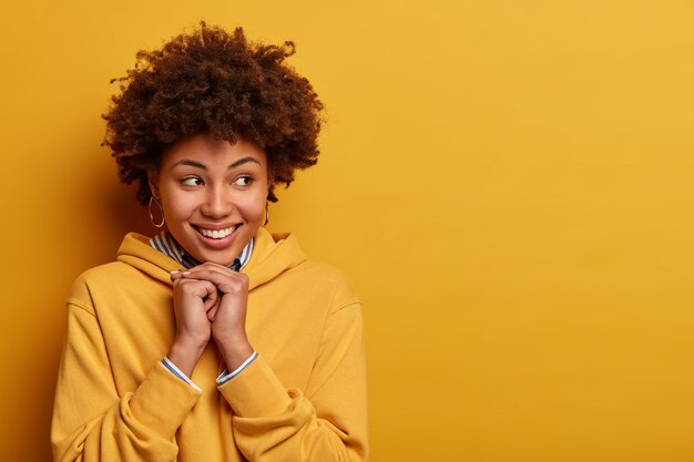 Portrait of pleasant looking woman with curly hair, looks gladfully aside, being in high spirit, dressed in sweatshirt, poses against yellow wall. Human face expressions and emotions concept