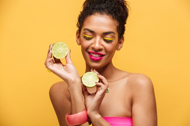 Portrait of pleased afro american woman with trendy makeup holding two halves of fresh lime in both hands isolated, over yellow wall