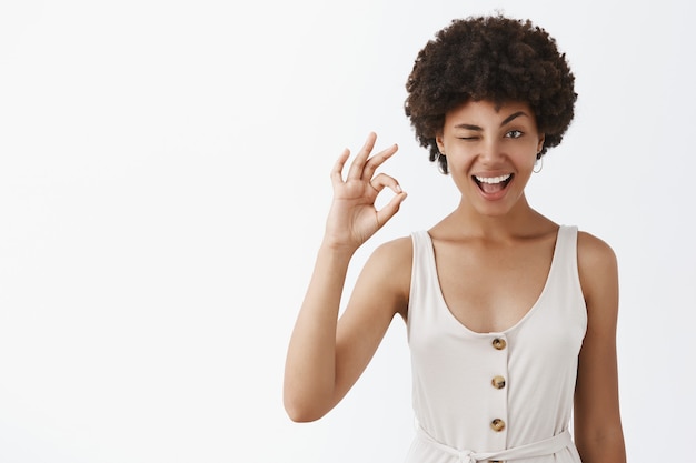 Portrait of pleased and confident emotive woman with dark skin and afro hairstyle winking with hint and smiling while showing okay gesture