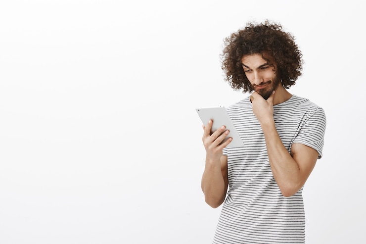 Free Photo portrait of pleased curious attractive guy with beard and afro haircut, rubbing chin while gazing at digital tablet, thinking or considering