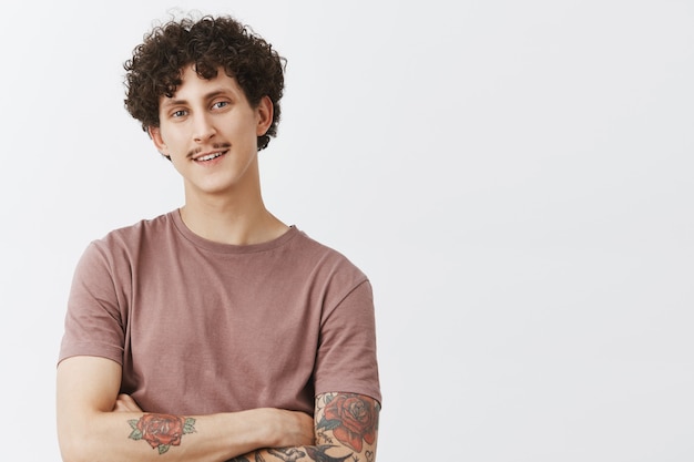 Portrait of pleased and delighted handsome young guy with moustache tattoos and curly hairstyle smiling from happy and satisfied feelings