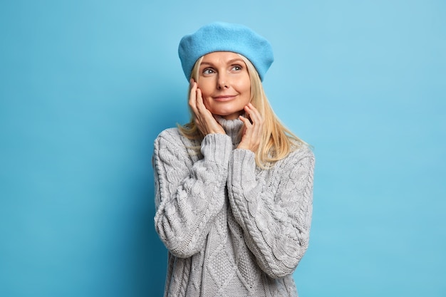 Portrait of pleased dreamy woman touches face gently looks away with peaceful expression wears beret grey knitted sweater.