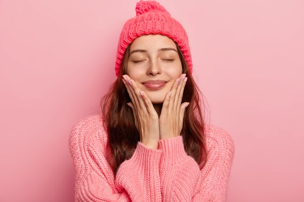 Portrait of pleased European woman touches both cheeks with palms, wears knitted hat and sweater, closes eyes with satisfaction