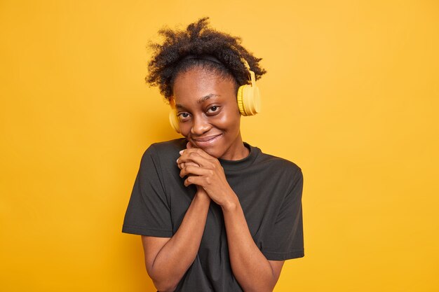 Portrait of pleased good looking Afro American teenage girl feels shy keeps hands near face looks satisfied at camera