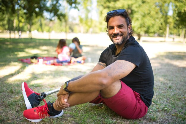 Portrait of pleased man with disability on picnic in park. Dark-haired man in shorts and black t-shirt posing, sitting on grass. Children in background. Disability, family, love concept