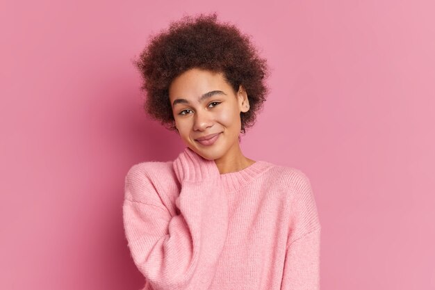 Portrait of pleased millennial woman keeps hand on neck looks gently and wears casual jumper