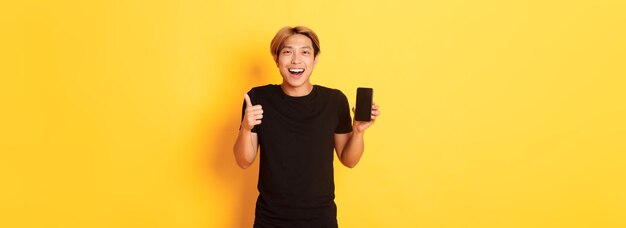 Portrait of pleased smiling asian guy showing smartphone screen and thumbsup satisfied with applicat