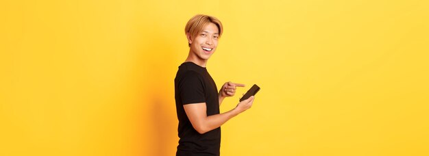 プロファイルに立って、スマートフォンで指を指している満足のいく笑顔のハンサムなアジア人の肖像画