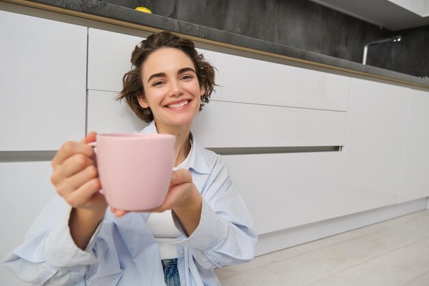 기하고 미소 짓는 젊은 여성의 초상화는 그녀의 음료를 추천합니다. 핑크색 차 또는 커피 컵을 보여줍니다.