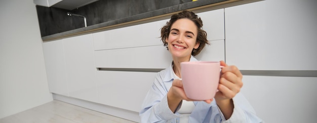 Free photo portrait of pleased smiling young woman recommends her drink shows pink cup of tea or coffee with