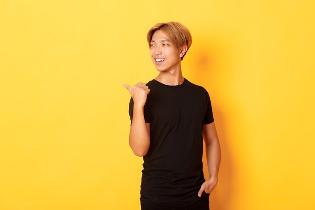 Portrait of pleased thoughtful asian guy, looking and pointing finger left at logo, standing yellow wall
