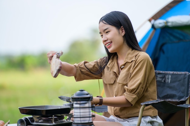 혼자 피크닉 팬에 구운 돼지고기 바베큐를 하고 캠핑장의 의자에 앉아 음식을 요리하는 행복한 젊은 아시아 여성의 초상화