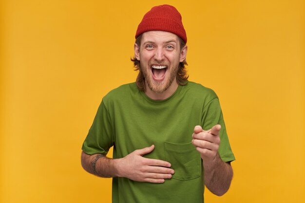 Portrait of positive, adult male with blond hairstyle and beard. wearing green t-shirt and red beanie. has tattoo. laughing hard from you. isolated over yellow wall