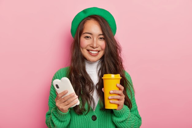 Portrait of positive brunette girl has coffee break after lectures, uses modern mobile phone for viewing pictures in social networks, sends notification to friend