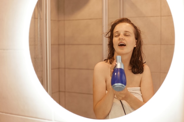 Portrait of positive and cheerful young adult beautiful woman with dark hair and a towel on her body blowing to her face with hair dryer and singing, morning routine.