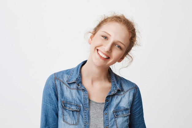 Portrait of positive friendly european redhead girl tilting head right and smiling broadly, glancing at camera with pure blue eyes