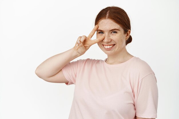 Portrait of positive ginger girl shows her v-sign, peace gesture near face and smiling happy, cheerful mood, showing perfect no makeup facial skin with natural freckles, white background.