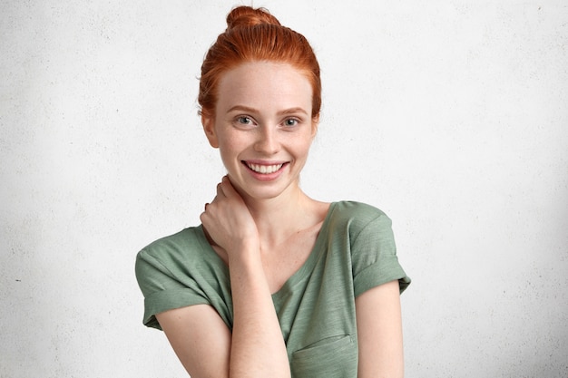 Free photo portrait of positive ginger young female model being satisfied to be photographed, has broad smile and white teeth, poses against concrete wall.