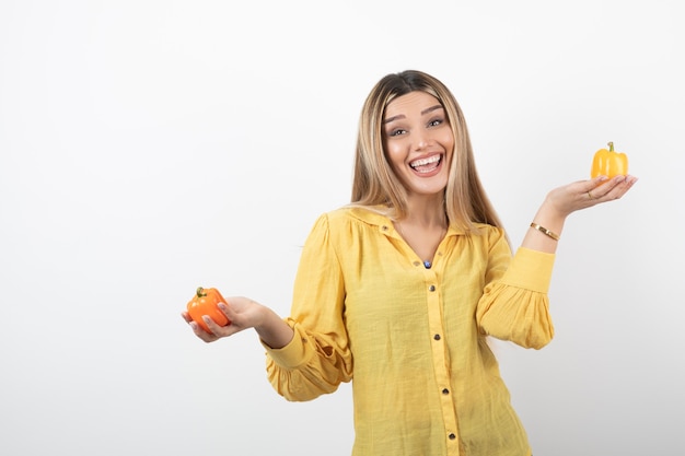 Portrait of positive girl holding colorful bell peppers on white wall.