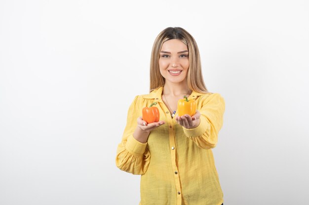 Portrait of positive girl offering colorful bell peppers on white wall.