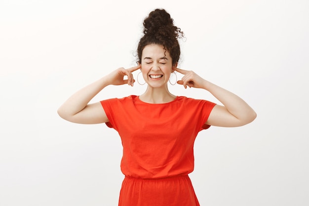 Portrait of positive good-looking woman in red dress, smiling broadly and covering ears with index fingers