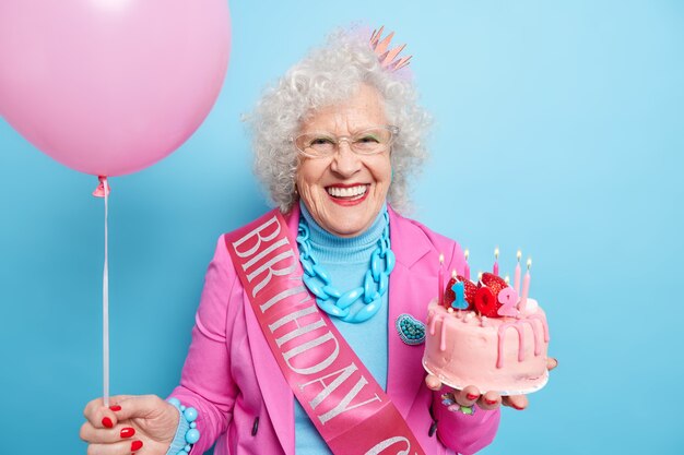 긍정적 인 회색 머리 여자의 초상화 102 번째 생일을 축하하고 맛있는 케이크와 팽창 된 풍선을 보유하고 있습니다.