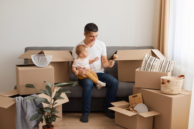 Portrait of positive optimistic man wearing white t-shirt and jeans sitting on sofa with his baby daughter, moving to a new flat, posing indoor, playing with his infant kid.