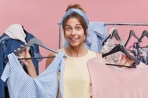 Portrait of positive pretty woman holding hangers with clothes choosing between two gorgeous dresses, waiting for your advice. Female shopaholic enjoying shopping in the sale, buying new garment