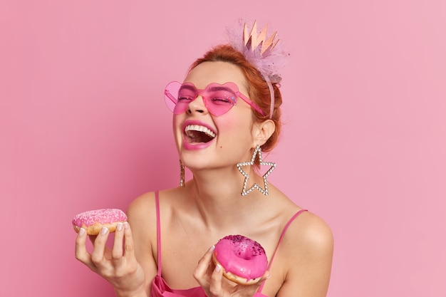 Портрет позитивной рыжей кавказской женщины громко смеется, держит рот открытым, в приподнятом настроении держит два пончика