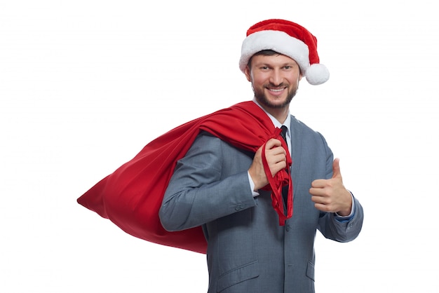 Ritratto di babbo natale positivo nella suite grigia, berretto rosso e borsa piena sulla spalla, sorridendo e mostrando super.