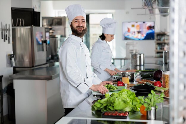 Portrait of positive smiling head chef standing in professional restaurant kitchen while looking at camera. Confident man wearing food industry white uniform while preparing delicious meal.