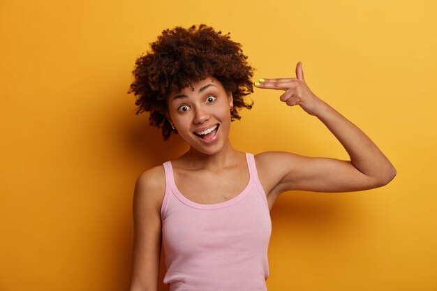 Portrait of positive teenage girl makes suicide gesture, shoots in temple with fingers, gazes happily , wears casual t shirt, feels bored, tries to amuse herself, stays at home, isolated