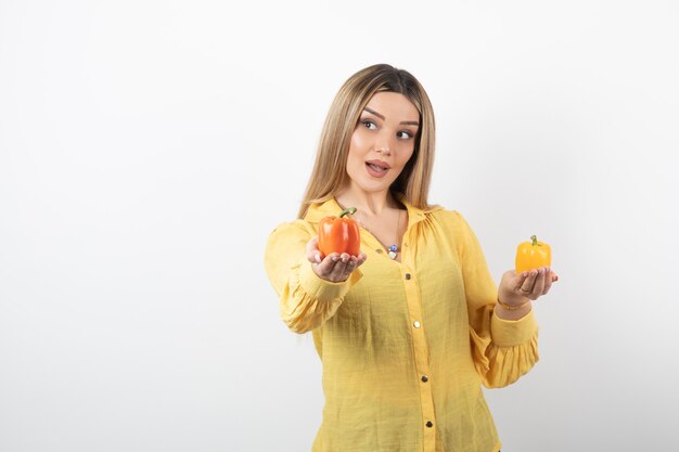 Portrait of positive woman offering colorful bell peppers over white wall.