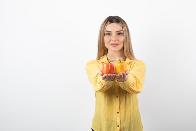 Portrait of positive woman offering colorful bell peppers over white wall.